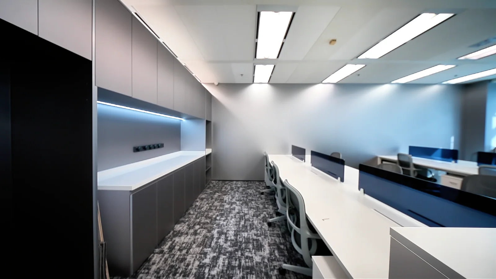 Grey built-in cabinets with linear lighting beside white desks and blue divider screens.