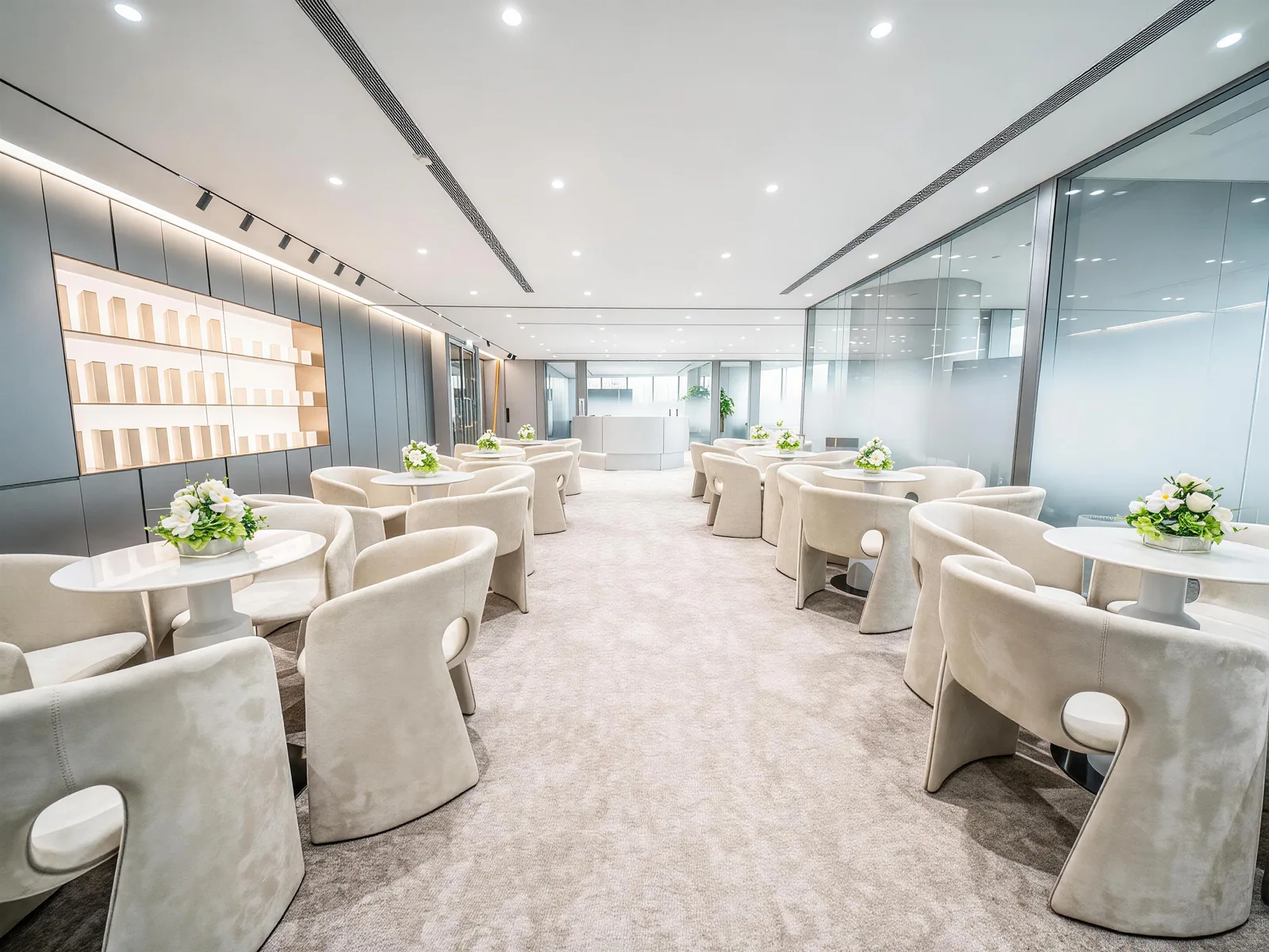 A central aisle between pale lounge chairs and round tables, with illuminated shelving and glazed rooms in the Wellness Brand Office.